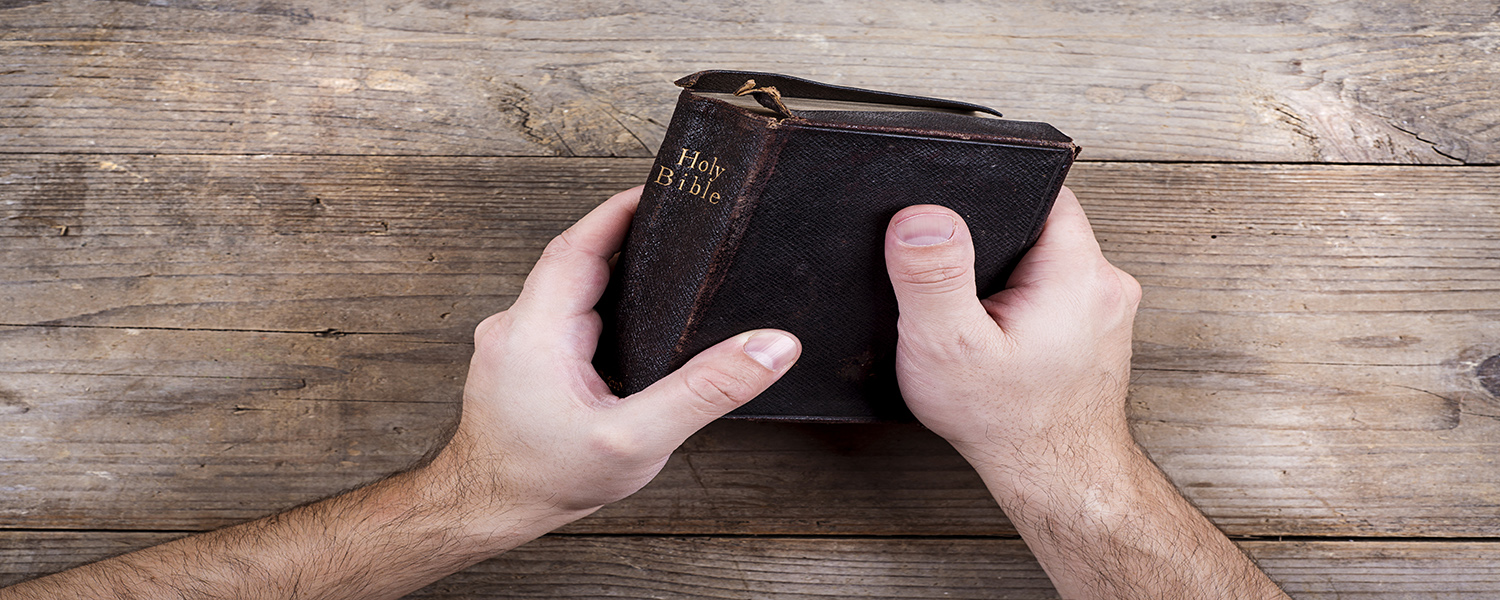 Hands holding Bible on a wooden desk background.